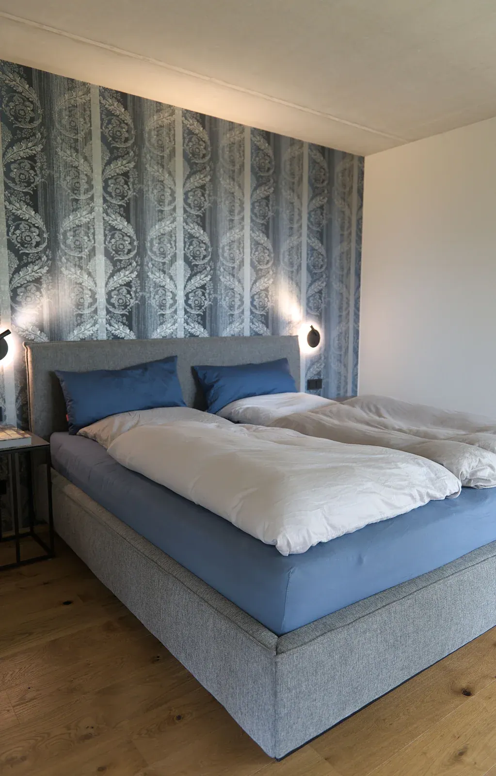 Schlafzimmer mit blau-grauem Polsterbett, gemusterter Tapete und Wandleuchten