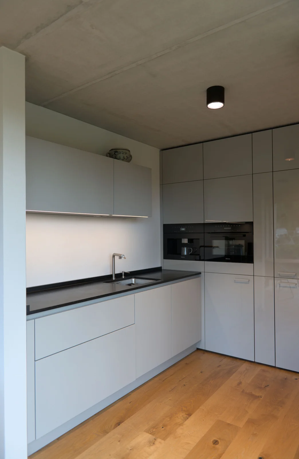 Küche mit hellgrauen Fronten, schwarzer Arbeitsplatte, Sichtbetondecke und Holzboden