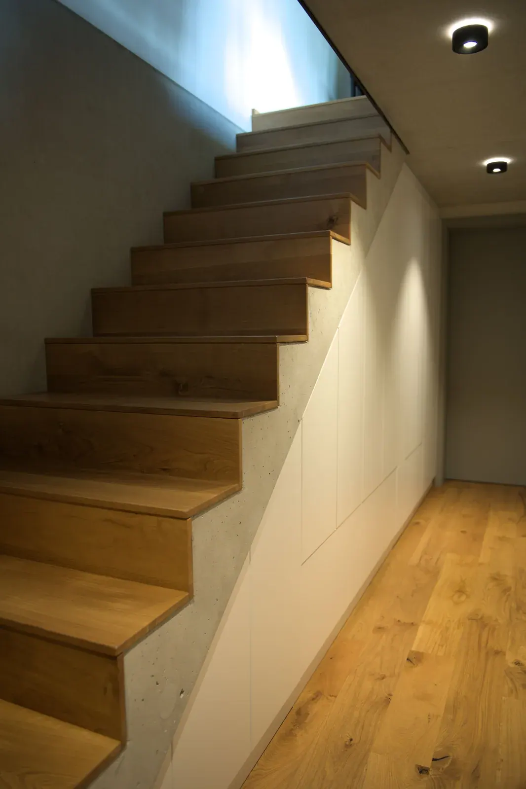 Holztreppe mit hellen Einbauschränken unter den Stufen und dunklen Deckenleuchten